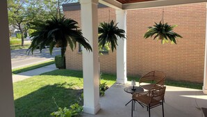 A covered patio with a brick wall, two hanging plants, a table with chairs, and a view of a street with trees and a house.