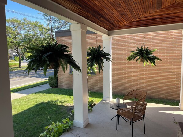 A covered patio with a brick wall, two hanging plants, a table with chairs, and a view of a street with trees and a house.