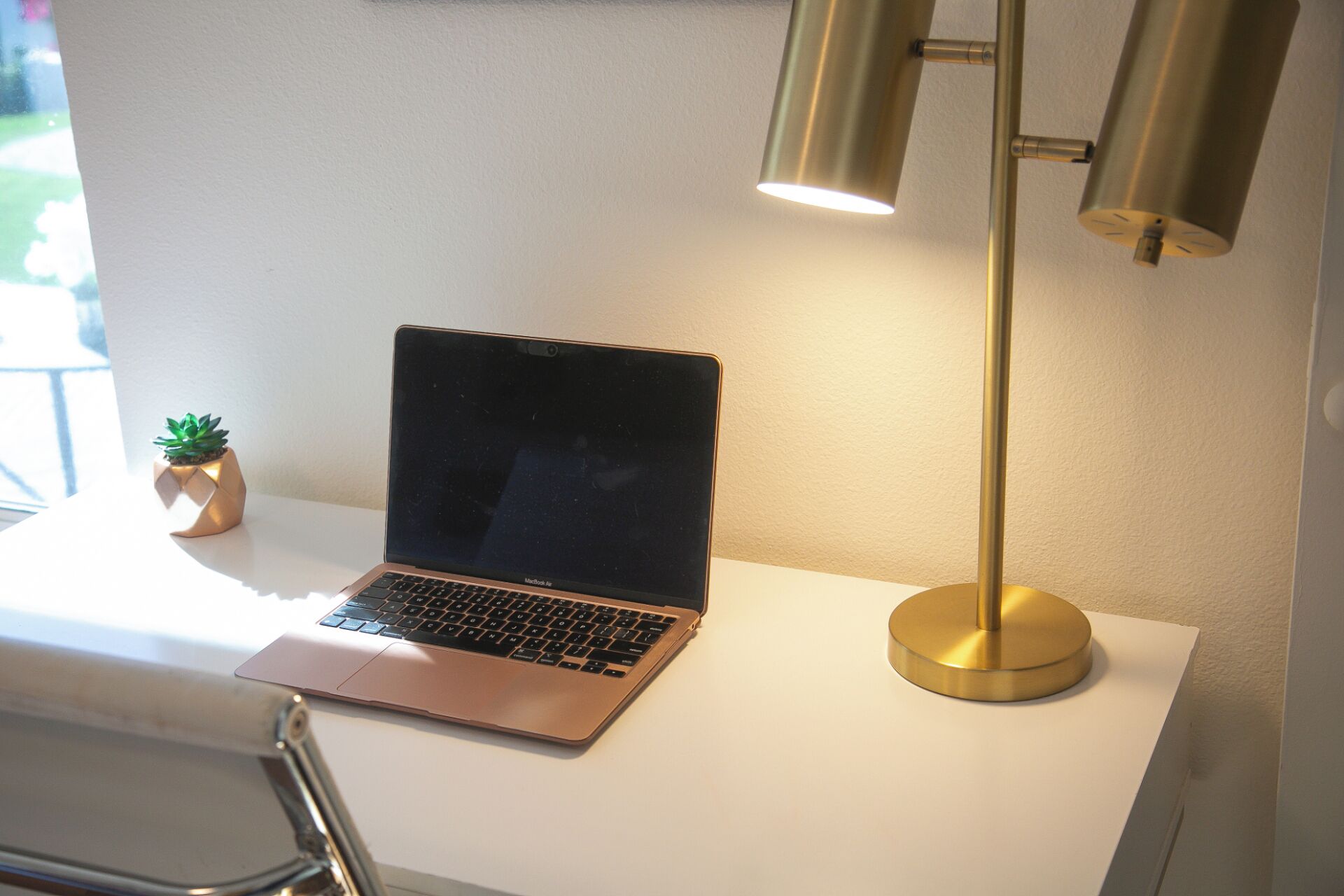 Un ordinateur portable sur un bureau blanc, avec une lampe dorée et une petite plante en pot.
