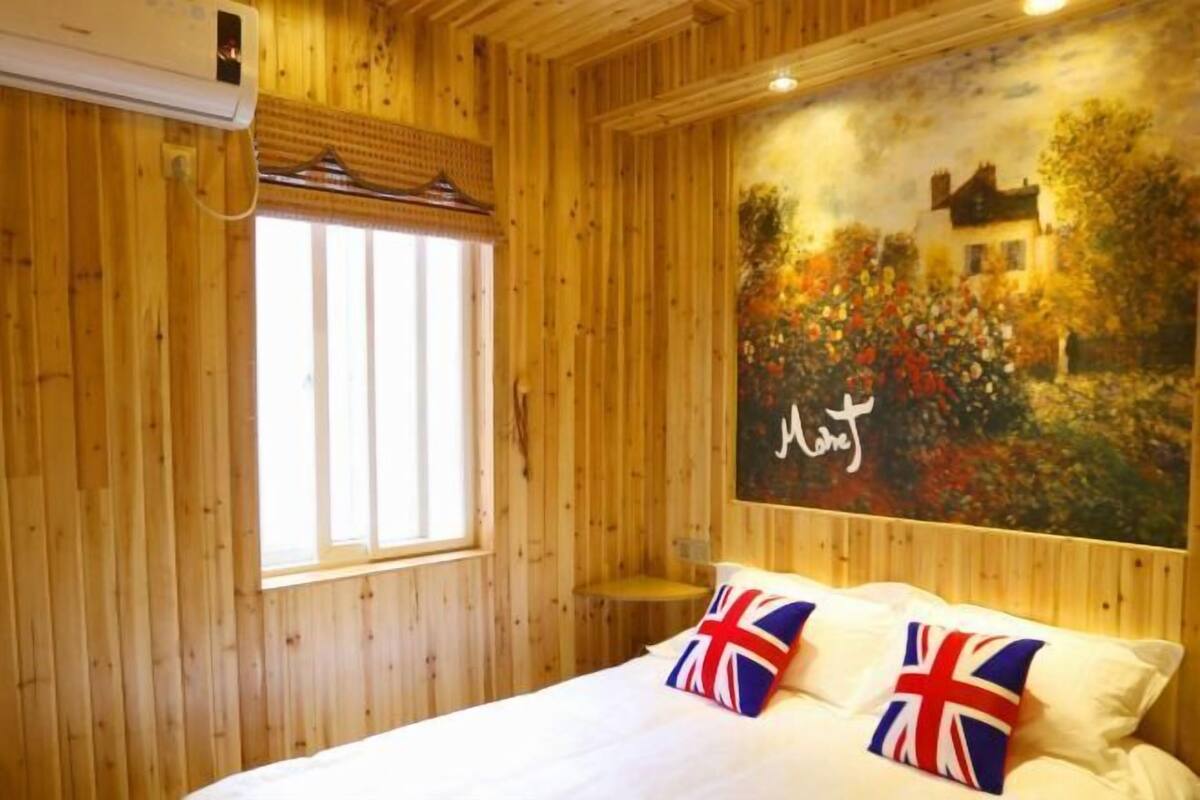 A wooden-paneled room with a bed, a window, and a painting of a house and garden.