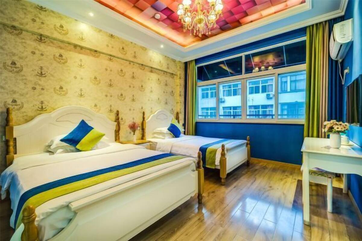 A hotel room with two beds, a chandelier, a window with a view of buildings, and a desk with a vase of flowers.