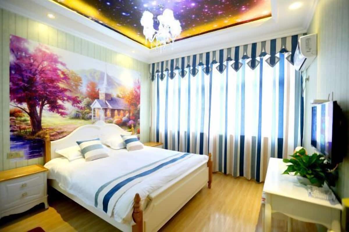 A bedroom with a large bed, a nightstand, a desk with a plant, and a mural on the wall.