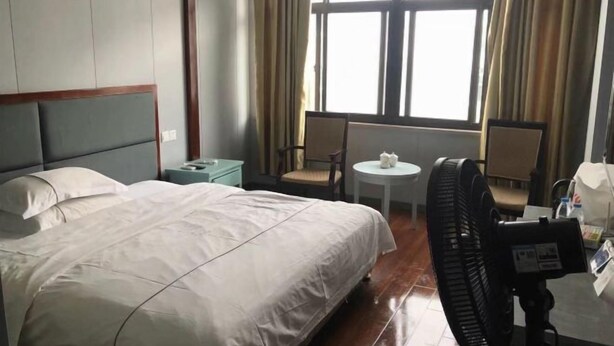 A hotel room with a bed, a small table, two chairs, a fan, and a window with curtains. - Hua Yang Hotel (Yuhuan)