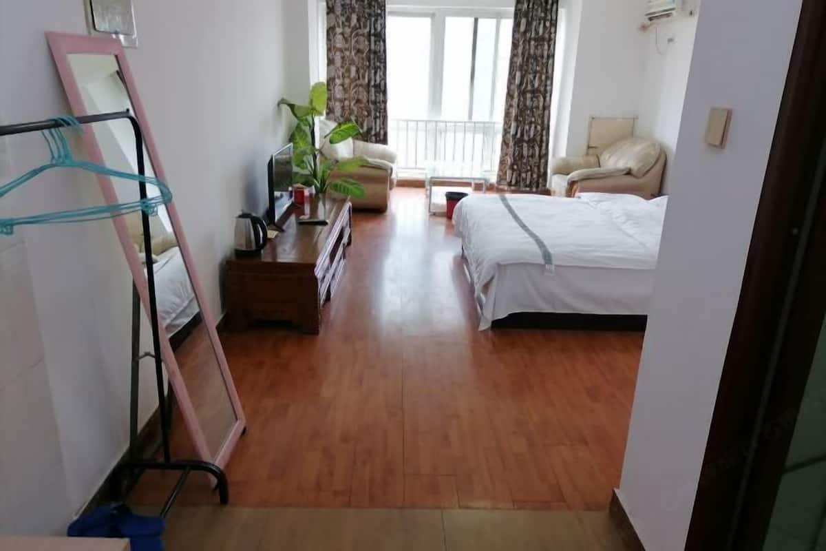 A hotel room with a bed, a chair, a desk with a plant, a mirror, and a window with curtains.