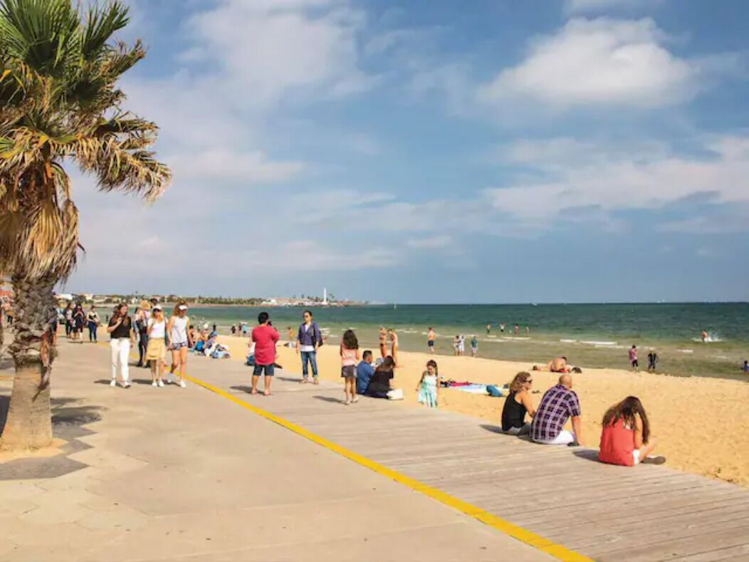 Une plage avec une promenade en bois, des palmiers, et des gens qui se détendent sur le sable et qui sont assis sur la promenade.