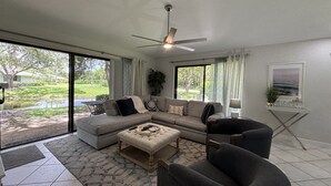 A spacious living room with a large sectional sofa, a round coffee table, and a dark armchair. There is a ceiling fan, a window with curtains, and a view of a garden.