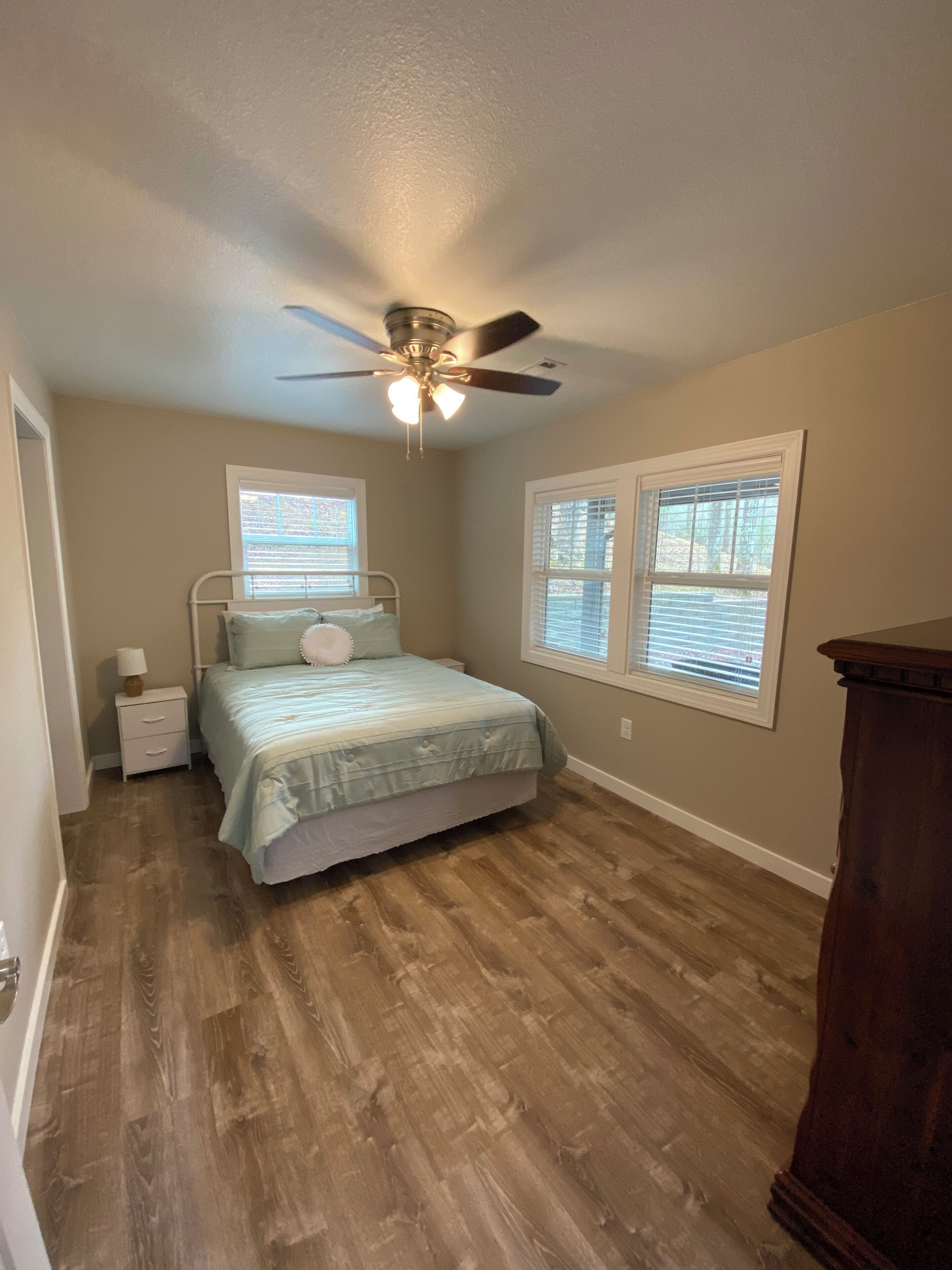 A bedroom with a bed, nightstand, ceiling fan, and two windows with blinds.
