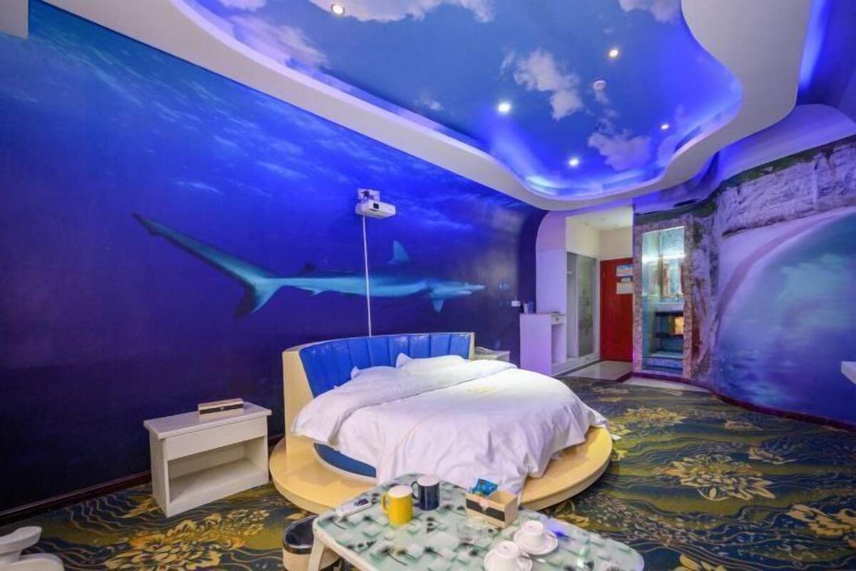 Una habitación con cama, una mesita con objetos, un mural en la pared de una escena submarina y un techo con diseño de nubes.
