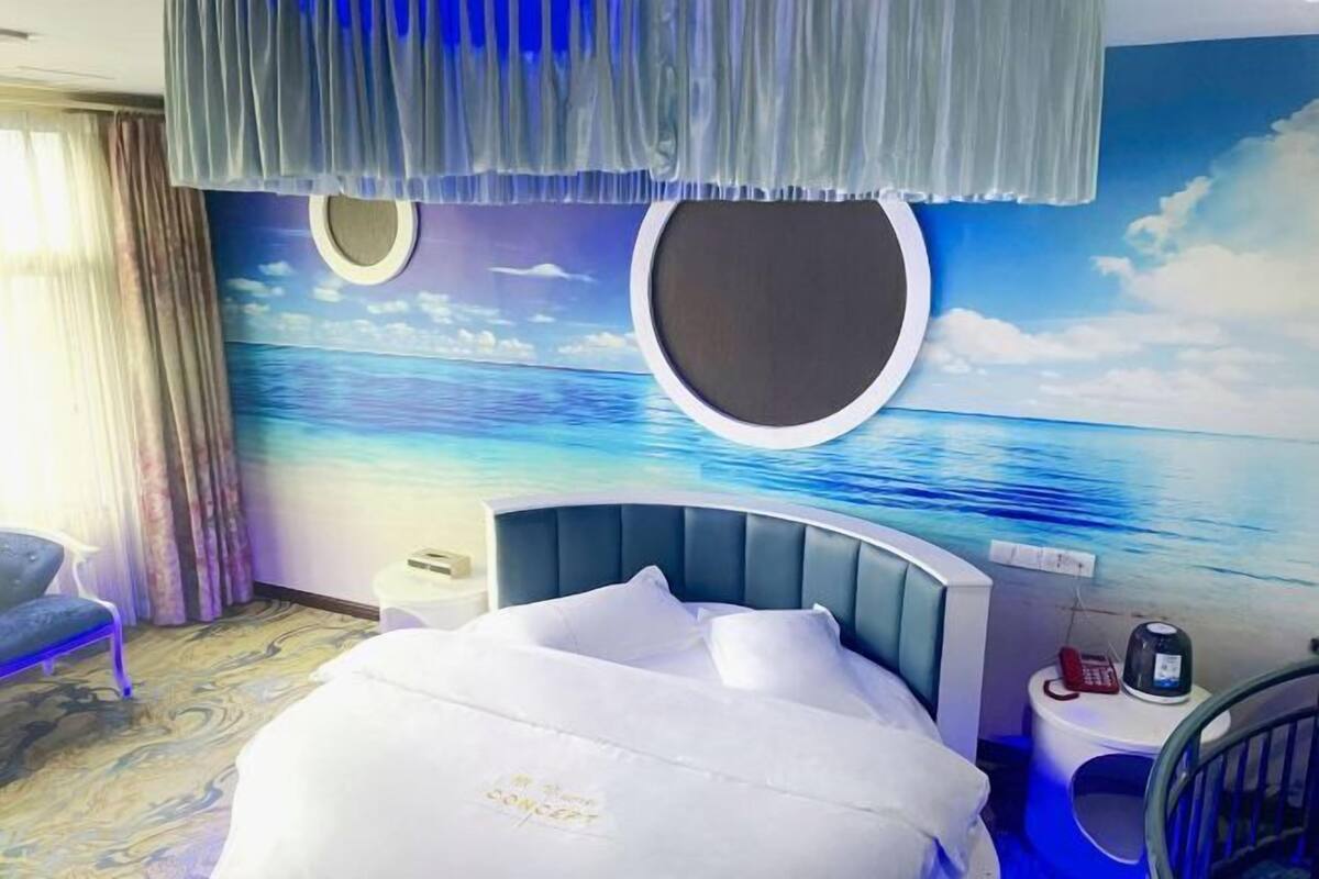 Habitación de hotel con cama, una silla, una mesita de noche y un mural en la pared que representa una playa.