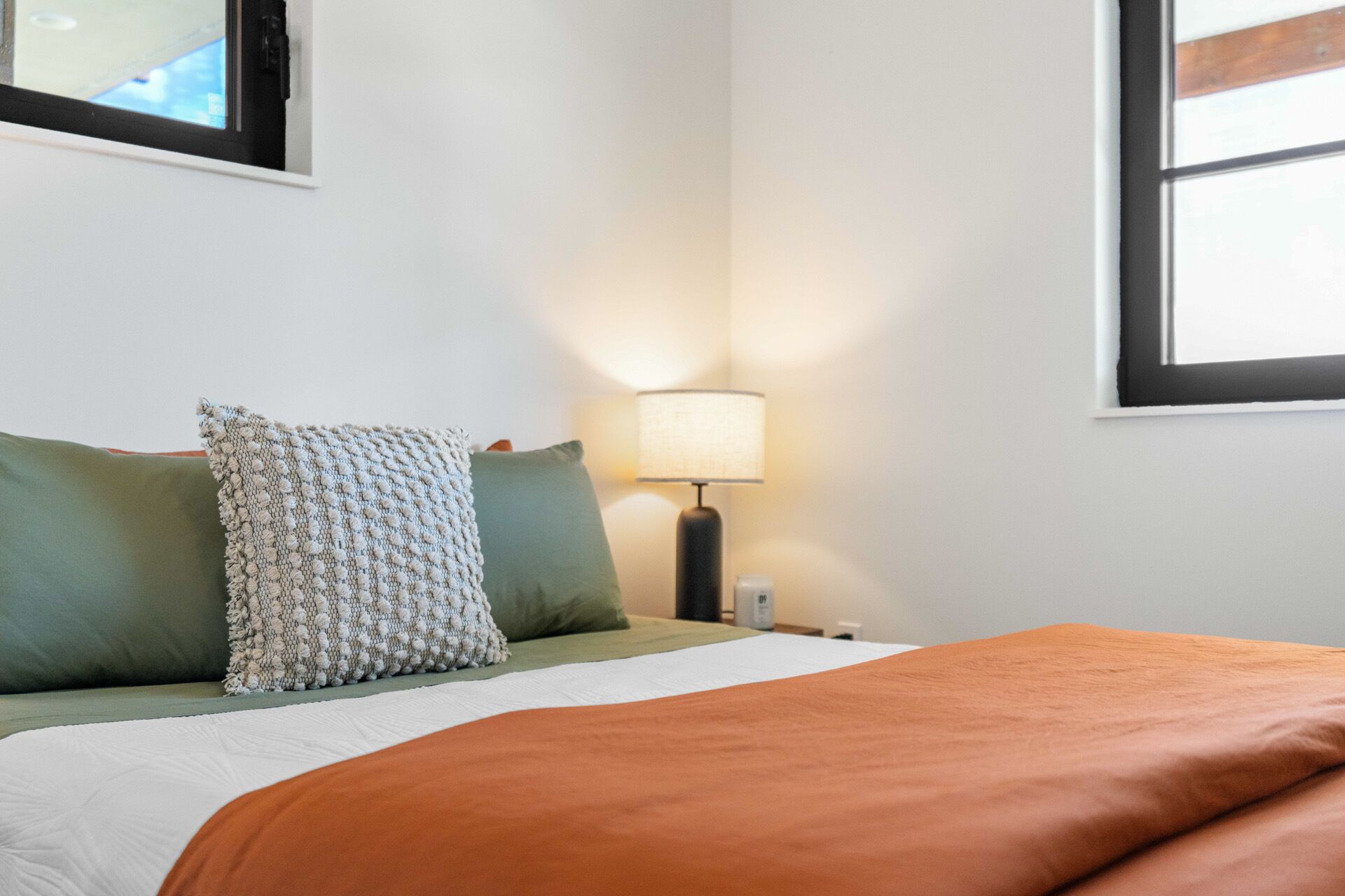 Una cama bien hecha con una manta blanca y naranja, almohadas verdes y blancas, y una lámpara en una mesita de noche.