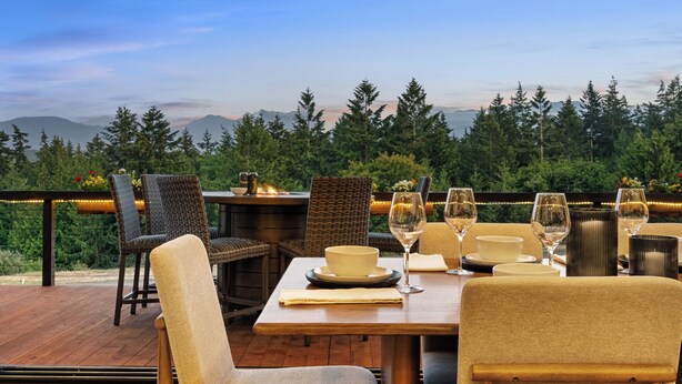 Un área de comedor al aire libre con sillas de mimbre y una mesa de madera preparada para una comida, con vista a una cadena montañosa boscosa.