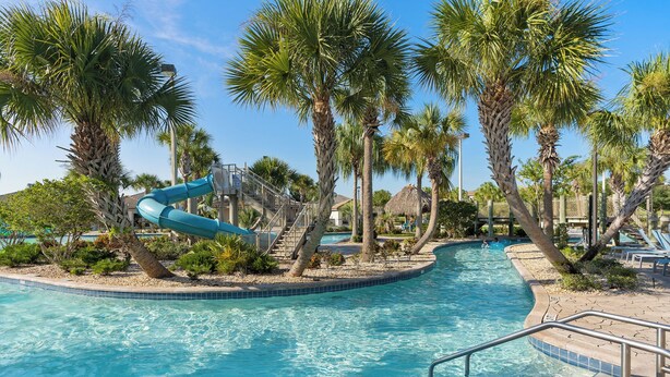 A swimming pool with a water slide, surrounded by palm trees and a thatched-roof structure. - Champions Gate Resort Home w BBQ Pool  (Davenport)