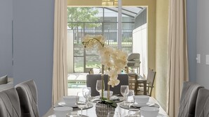 A dining room with a long table set for six, featuring white plates, bowls, and glasses, and a vase with white flowers. - Champions Gate Resort Home w BBQ Pool  (Davenport)