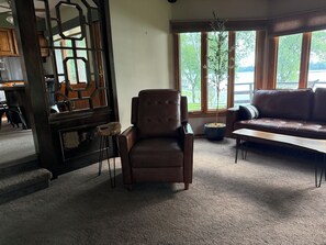 Un salon avec un fauteuil en cuir marron, un canapé, une table basse, une plante en pot et une vue sur l’extérieur grâce à de grandes fenêtres.