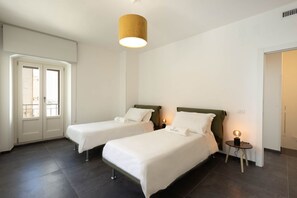 A hotel room with two single beds, a round table with a lamp, a door, and a window with a view.