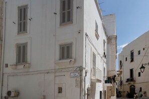 A white building with multiple windows and shutters, a sign indicating 'Largo S. Angelo,' and a narrow street with other buildings.