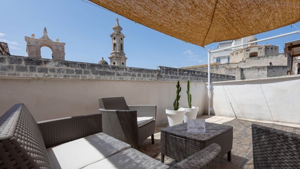 Une terrasse sur le toit, meublée de mobilier en osier, une enseigne indiquant «WePuglio», et une vue sur des bâtiments historiques en arrière-plan.