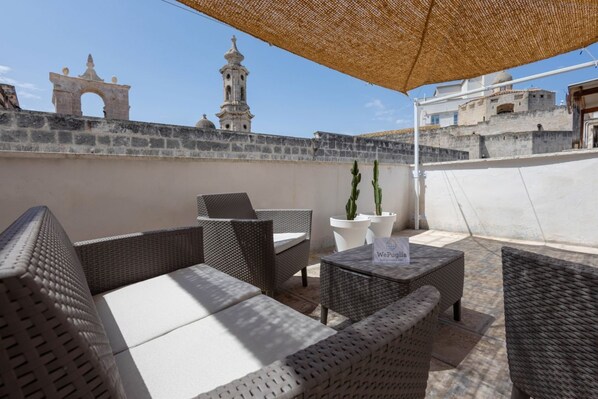 Eine Dachterrasse mit Korbmöbeln, einem Schild mit der Aufschrift 'WePuglio' und Blick auf historische Gebäude im Hintergrund.