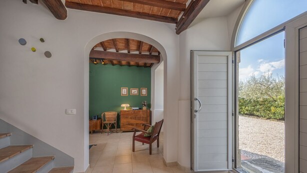 Un couloir avec un escalier en bois, une porte en arc donnant sur une pièce aux murs verts et ornée de tableaux encadrés, et une porte menant à un espace extérieur.