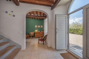 Un couloir avec un escalier en bois, une porte en arc donnant sur une pièce aux murs verts et ornée de tableaux encadrés, et une porte menant à un espace extérieur.