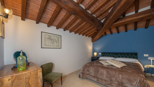A bedroom with a wooden ceiling, a bed with a brown bedspread, a chair, a desk with a green bottle, and a framed picture on the wall.