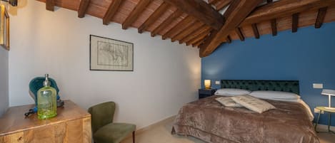 A bedroom with a wooden ceiling, a bed with a brown bedspread, a chair, a desk with a green bottle, and a framed picture on the wall.