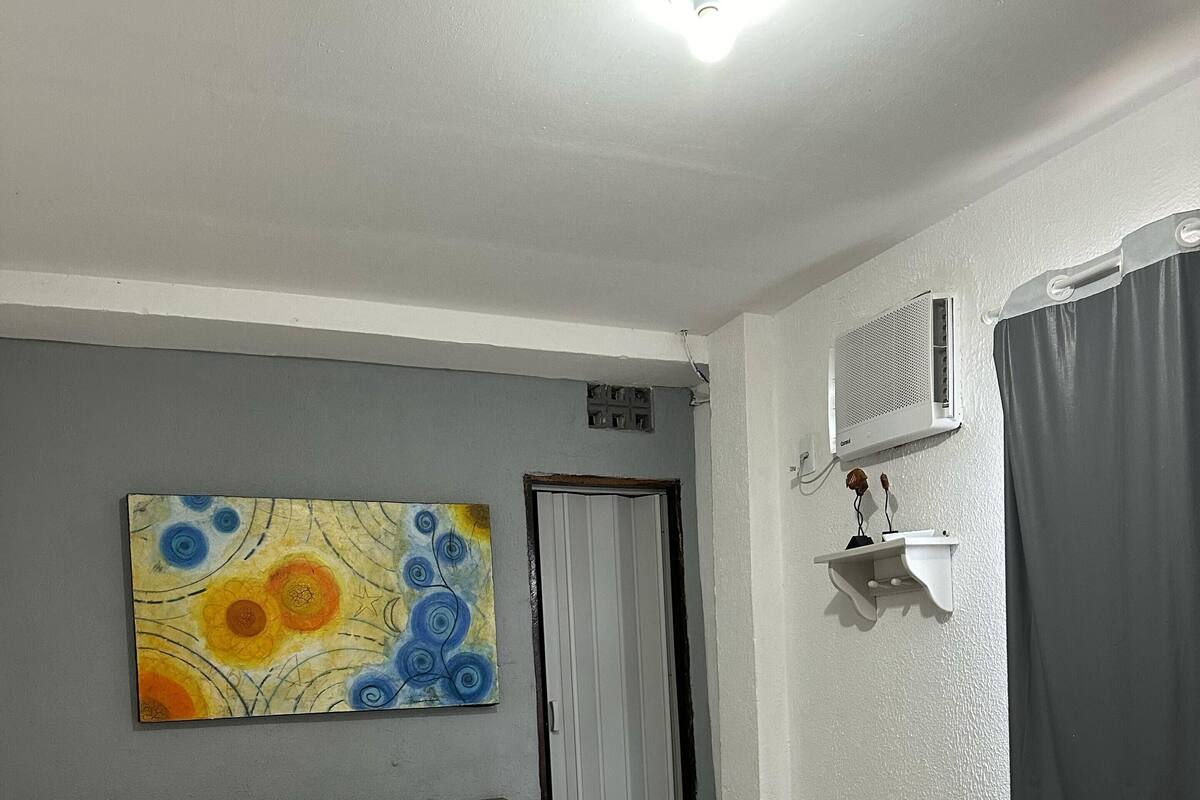Ett hotellrum med en säng, en tavla på väggen, ett fönster med gardiner och en väggmonterad luftkonditioneringsanläggning.