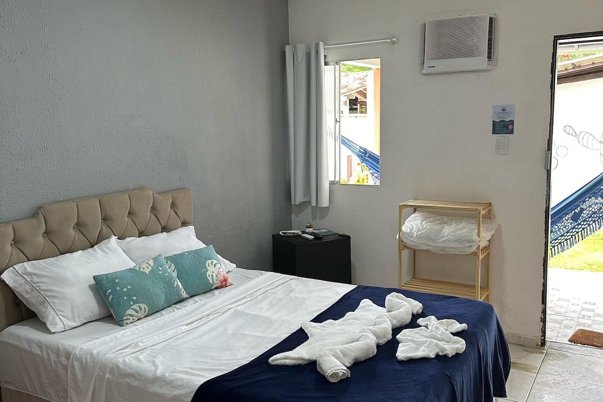 En snyggt bäddad säng med ett blått överkast, vita kuddar och en beige sänggavel. Bredvid sängen står ett litet sidobord med en fjärrkontroll, och på väggen sitter en luftkonditioneringsanläggning. Genom fönstret ser man en blå trappa och grönskande växter.