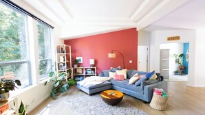 En moderne stue med blå sofa, et træbord og et gråt tæppe. Store vinduer, en reol og en plante.