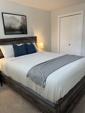 A bedroom with a bed, pillows, a blanket, a nightstand with a lamp, a door, and a framed picture on the wall. - The Haven at Pinecreek (Calgary)