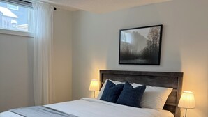 Ein ordentlich bezogenes Bett mit grauer Tagesdecke, zwei Nachttischlampen, ein gerahmtes Bild über dem Bett und ein Fenster mit Vorhängen.
