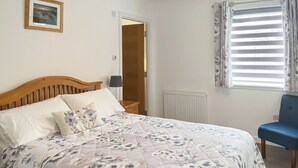 A bedroom with a wooden bed, floral bedding, a bedside table with a lamp, a chair, a window with curtains, and a framed picture on the wall.