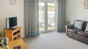 A living room with a television on a wooden stand, a sofa with cushions, a dining table with a vase and a remote control, and a large window with curtains.