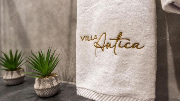 A white towel with "VILLA Antica" embroidered in gold, placed on a textured surface next to two small potted plants.