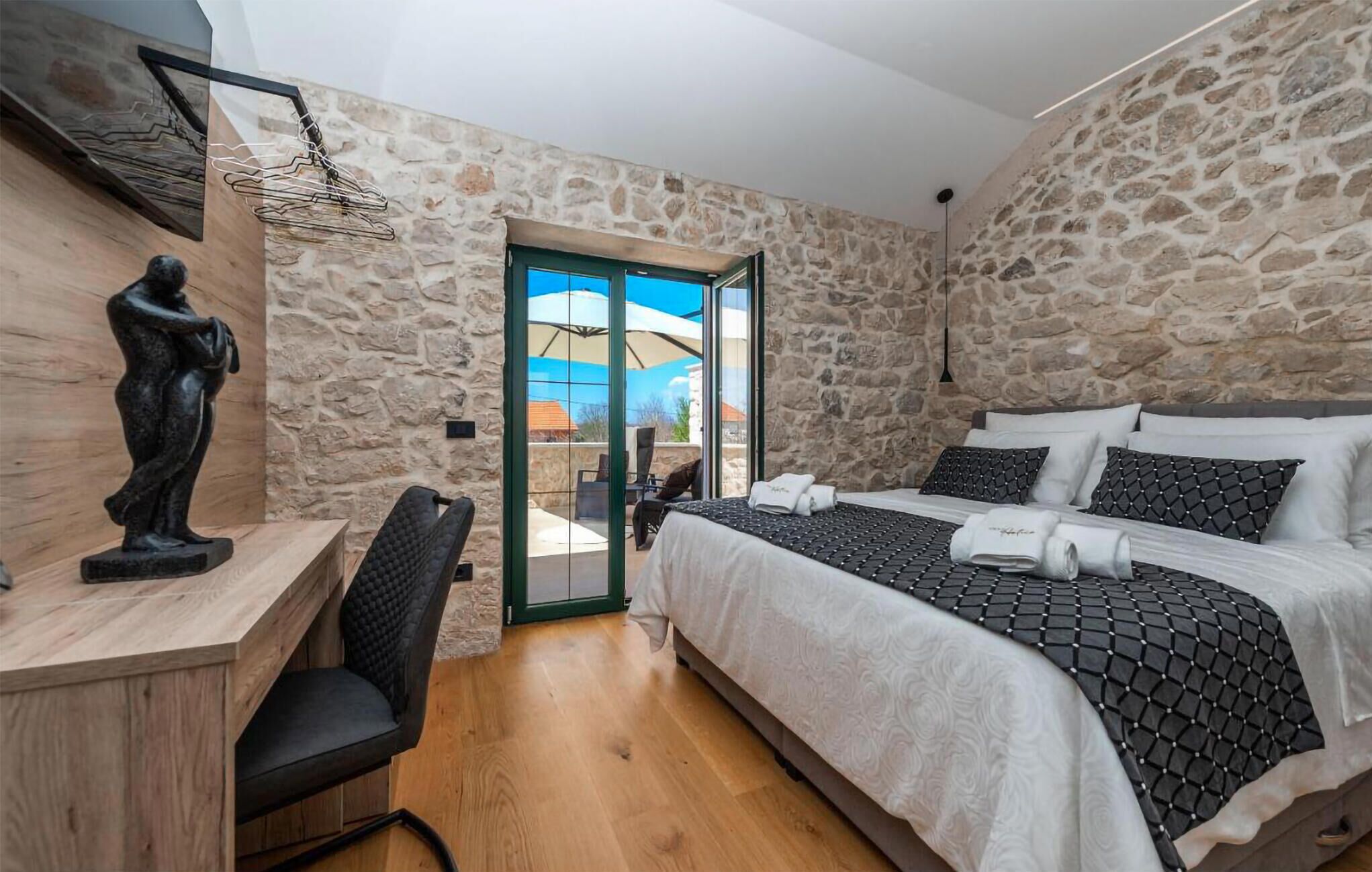 A bedroom with a stone wall, a bed with patterned bedding, a desk with a sculpture, and a view through a glass door.