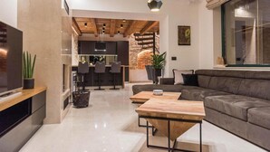 A modern living room with a grey sofa, wooden coffee table, and a kitchen area with a brick wall and wooden ceiling. - Luxurious home in Poljaci (Poljaci)