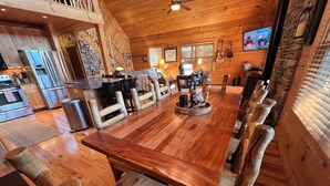 A wooden interior with a long dining table, chairs, a kitchen area, and a television mounted on the wall.