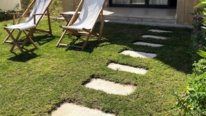 Un espacio exterior cuidado con dos sillones de descanso, un sendero de piedra y la entrada de un edificio con puerta corredera de vidrio.