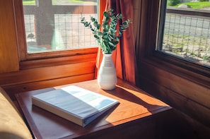 A wooden table with a book and a white vase with green plants by a window with orange curtains. - EML 7 Cloven-Hoofed (Paonia)