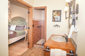 A bedroom with a wooden headboard, a bed with a patterned blanket, a small wooden stool, and a bathroom with a wooden sink and mirror. - EML 7 Cloven-Hoofed (Paonia)