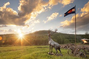 A deer statue, a flag, and a wagon wheel in a grassy field with a sunset in the background. - EML 7 Cloven-Hoofed (Paonia)