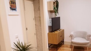 A living room with a wooden floor, a TV on a wooden stand, a white armchair, a potted plant, and a door with a framed picture. - Premium Apartment Porto Downtown, With Terrace (Porto)