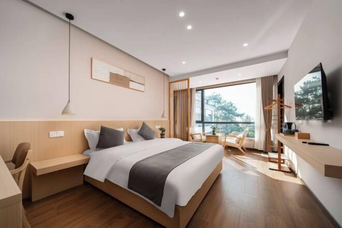 A modern hotel room with a large bed, a desk, a chair, a TV, and a balcony with a view.
