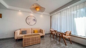 A modern living room with a sofa, coffee table, and chairs. There is a large window with sheer curtains, a hanging light fixture, and a wall-mounted artwork.