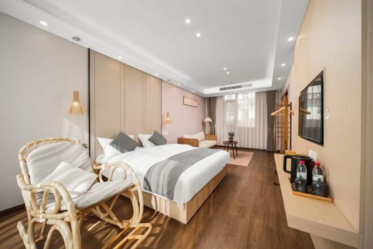 A modern hotel room with a large bed, a seating area with a chair, a small table, and a wall-mounted shelf with bottles.