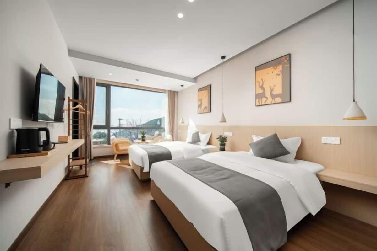 A modern hotel room with two beds, a TV, a desk with a coffee maker, and a large window.