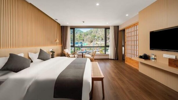 A hotel room with a large bed, a TV, a seating area with chairs, and a view of a mountainous landscape.