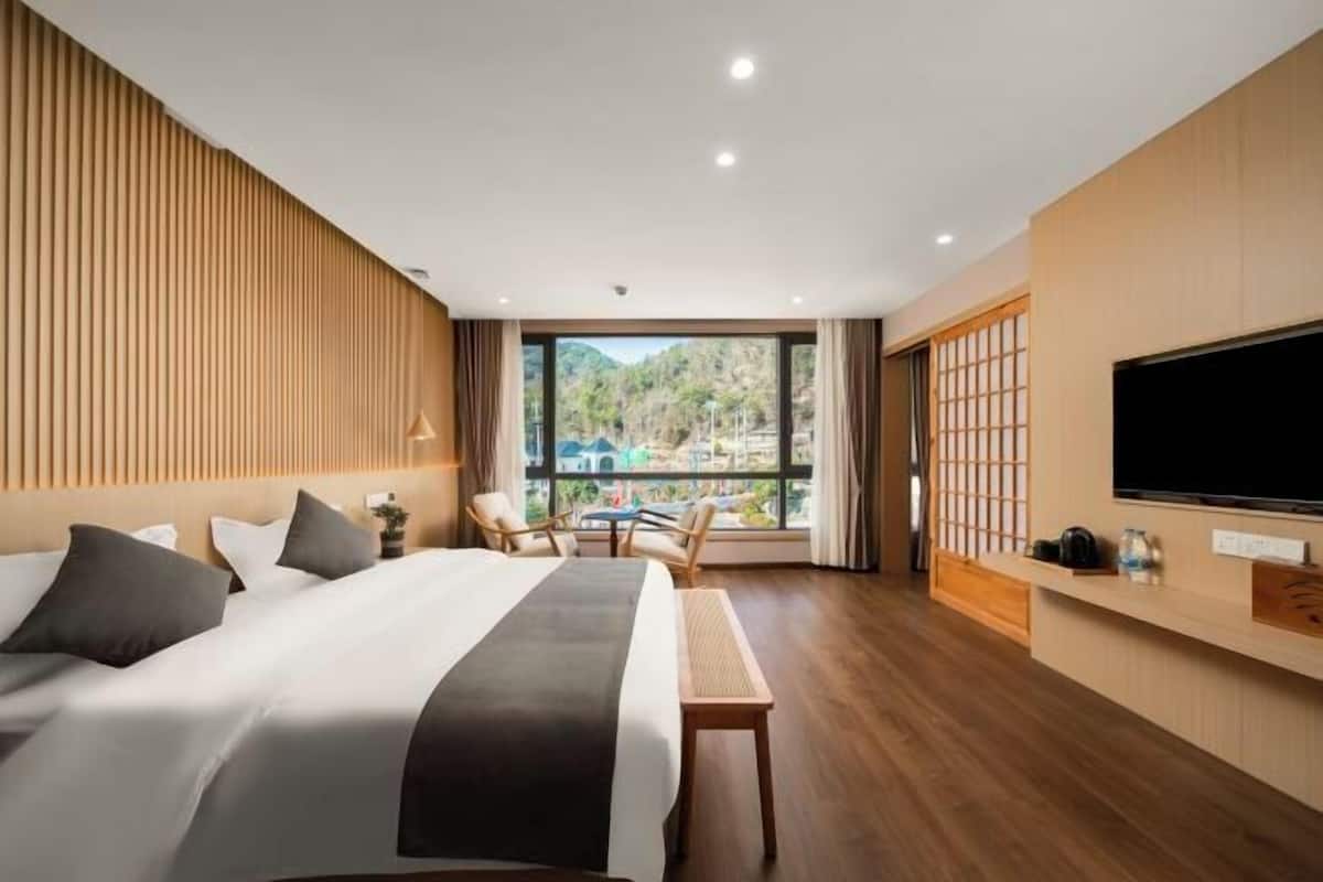 A hotel room with a large bed, a TV, a seating area with chairs, and a view of a mountainous landscape.