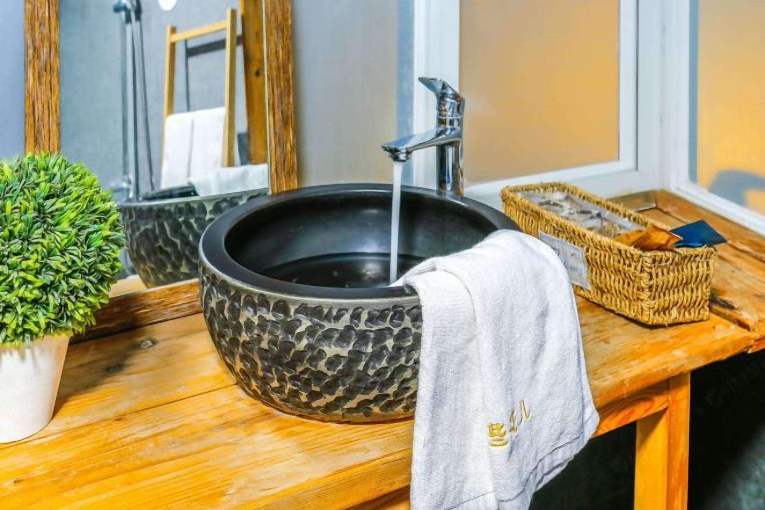 Un baño con un lavamanos de piedra negra, un mueble de tocador de madera, una pequeña planta en maceta y una cesta tejida.