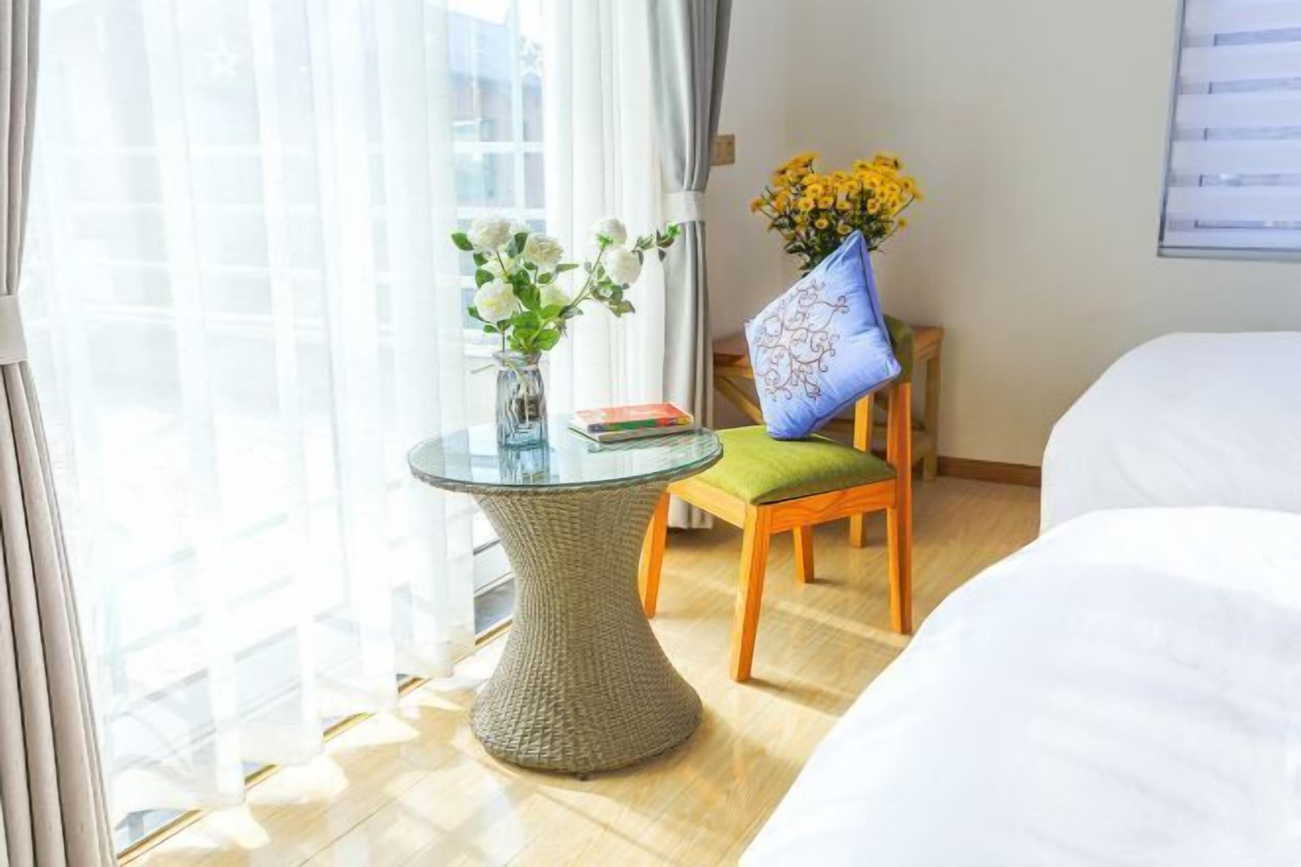 A hotel room with a bed, a small round table with a vase of flowers, a chair with a cushion, and a window with curtains.
